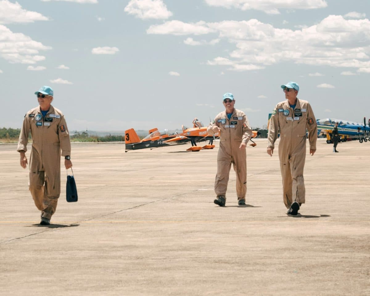 Three flight crew members walking across the tarmac past aerobatic aircraft
